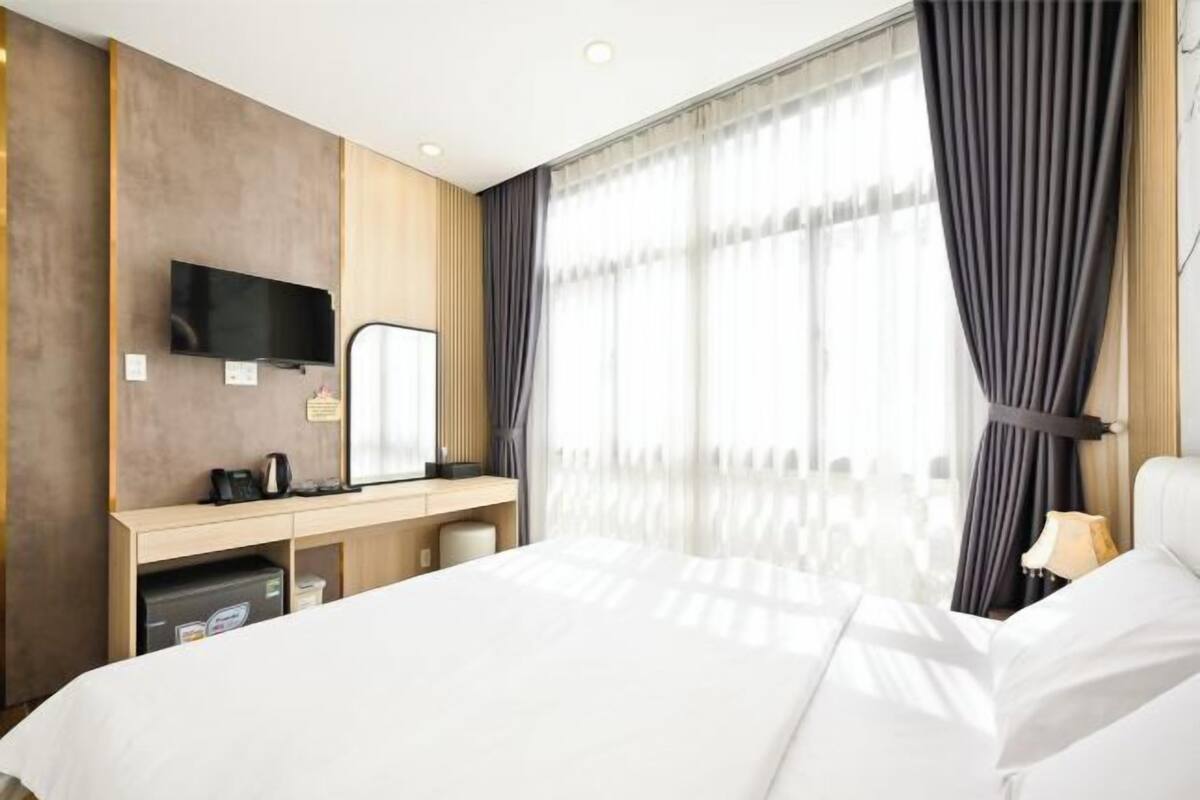 A hotel room with a bed, a desk with a flat-screen TV, a mirror, a lamp, and a window with curtains.