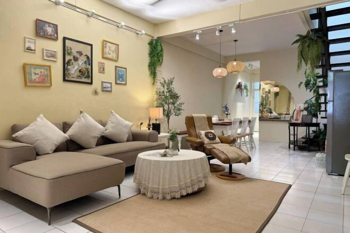 Uma sala de estar moderna com sofá, poltrona e mesa de centro redonda. Há quadros nas paredes e uma área de cozinha ao fundo.