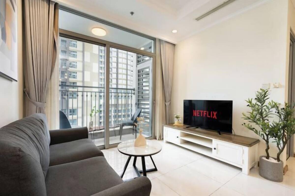 A modern living room with a grey sofa, a round coffee table, a TV on a white cabinet, and a balcony with a view of high-rise buildings.