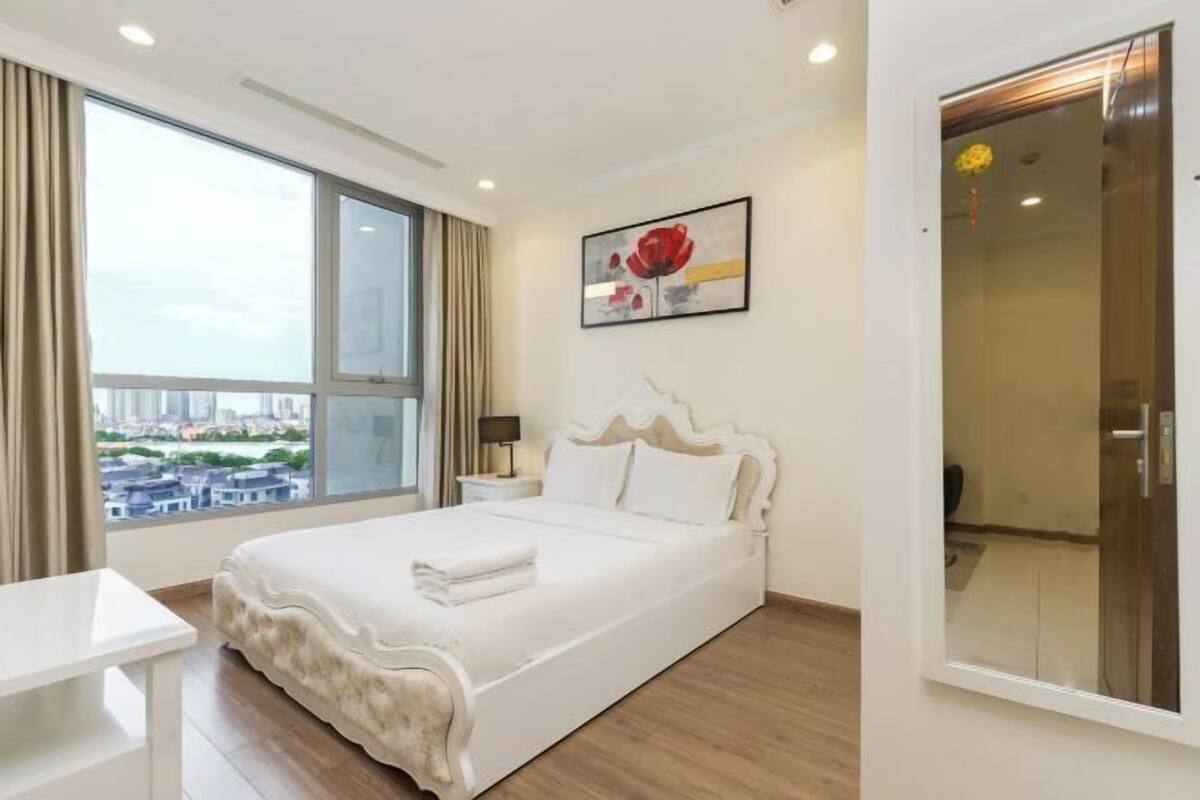 A hotel room with a large window, a bed, a bedside table, and a painting on the wall.
