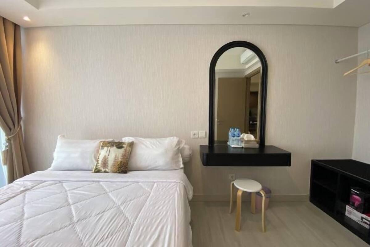 Une chambre d’hôtel avec un lit, un petit tabouret, un miroir et une étagère noire.