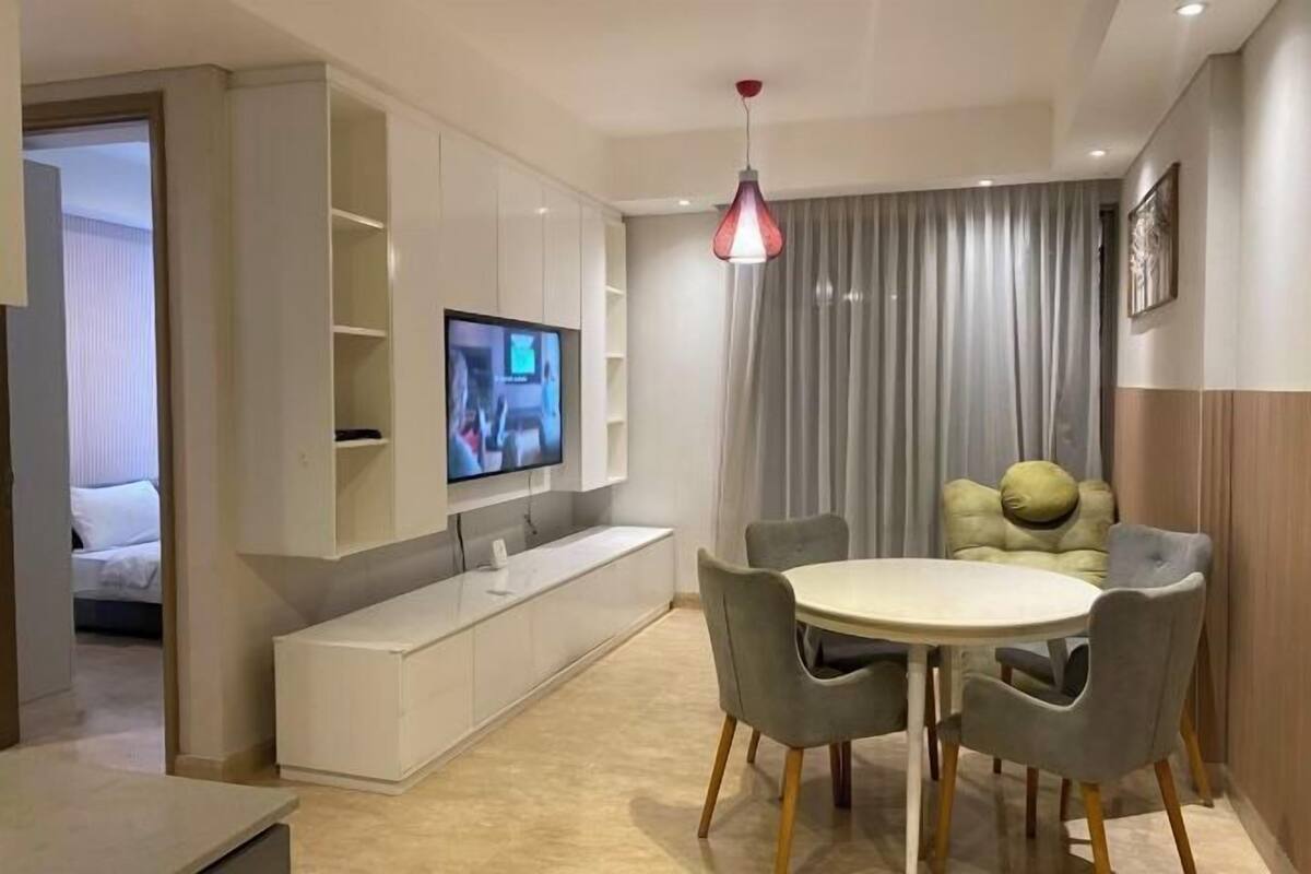Un salon moderne avec un téléviseur fixé au mur, une table de salle à manger ronde entourée de chaises et une grande fenêtre agrémentée de rideaux.