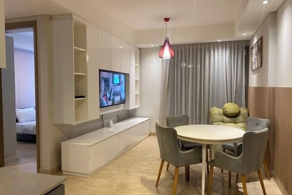 Un salon moderne avec un téléviseur fixé au mur, une table de salle à manger ronde entourée de chaises et une grande fenêtre agrémentée de rideaux.