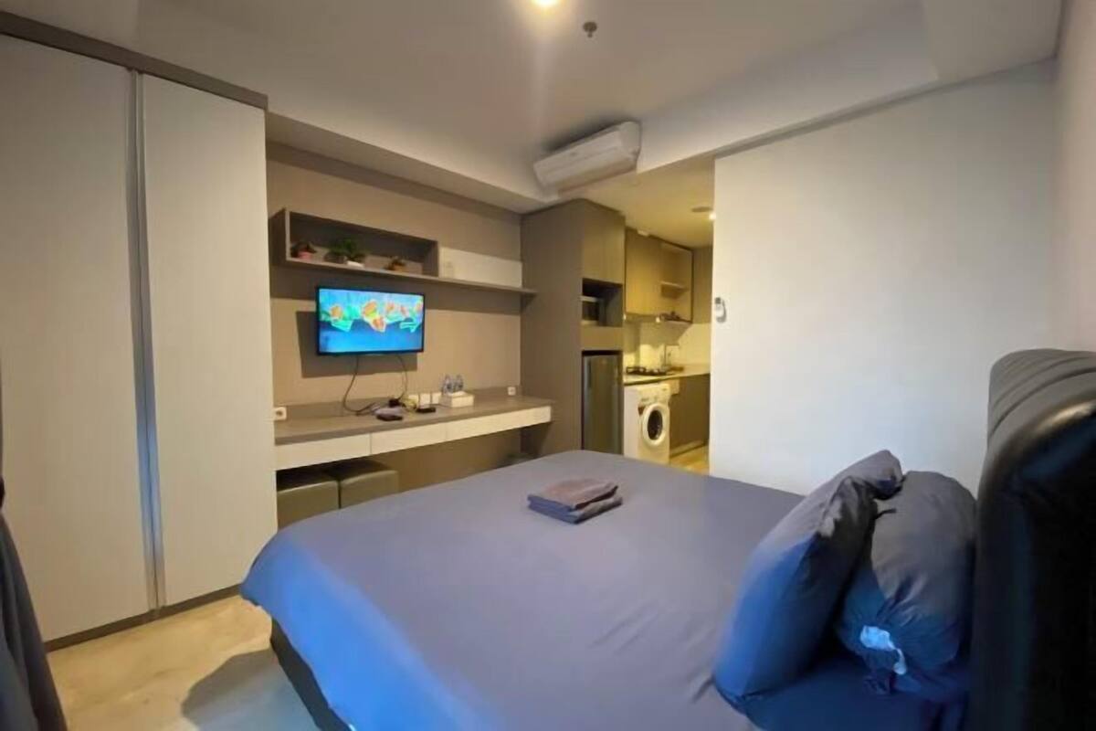 Une chambre à coucher moderne avec un lit, une télévision, un bureau et une machine à laver en arrière-plan.
