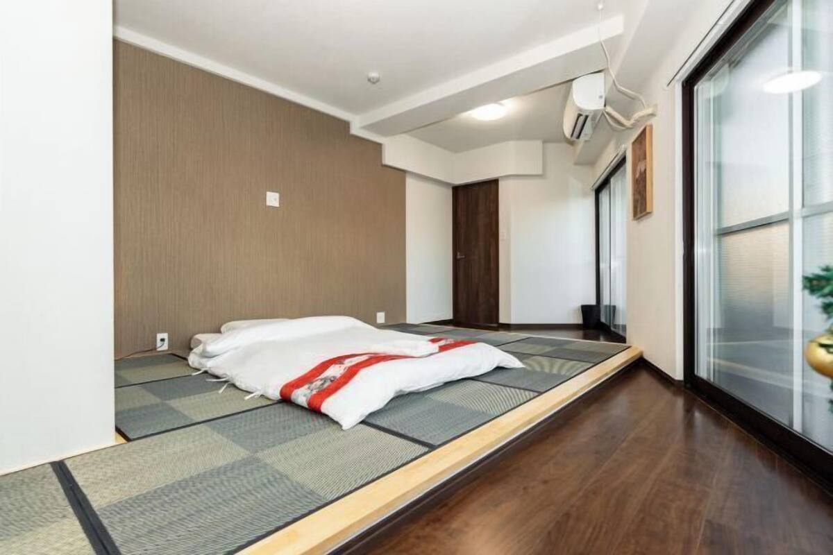 Ein Zimmer mit Tatami-Matten, einem Bett mit weißer und roter Decke, einer Holztür und einem Fenster mit Rollläden.