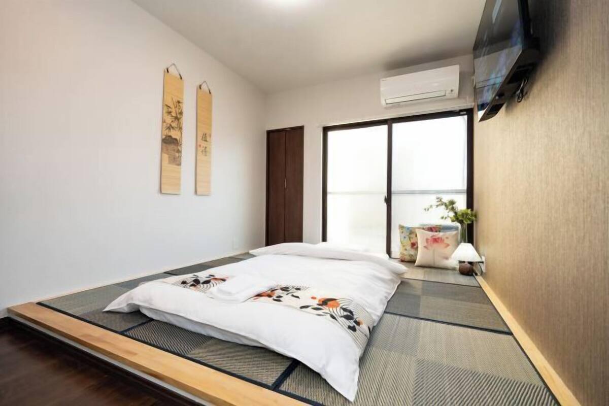 Ein schlicht eingerichteter Raum mit Tatami-Boden, einem Bett mit floralem Überwurf, einem an der Wand montierten Fernseher und einer Schiebetür, die zu einem Balkon führt.