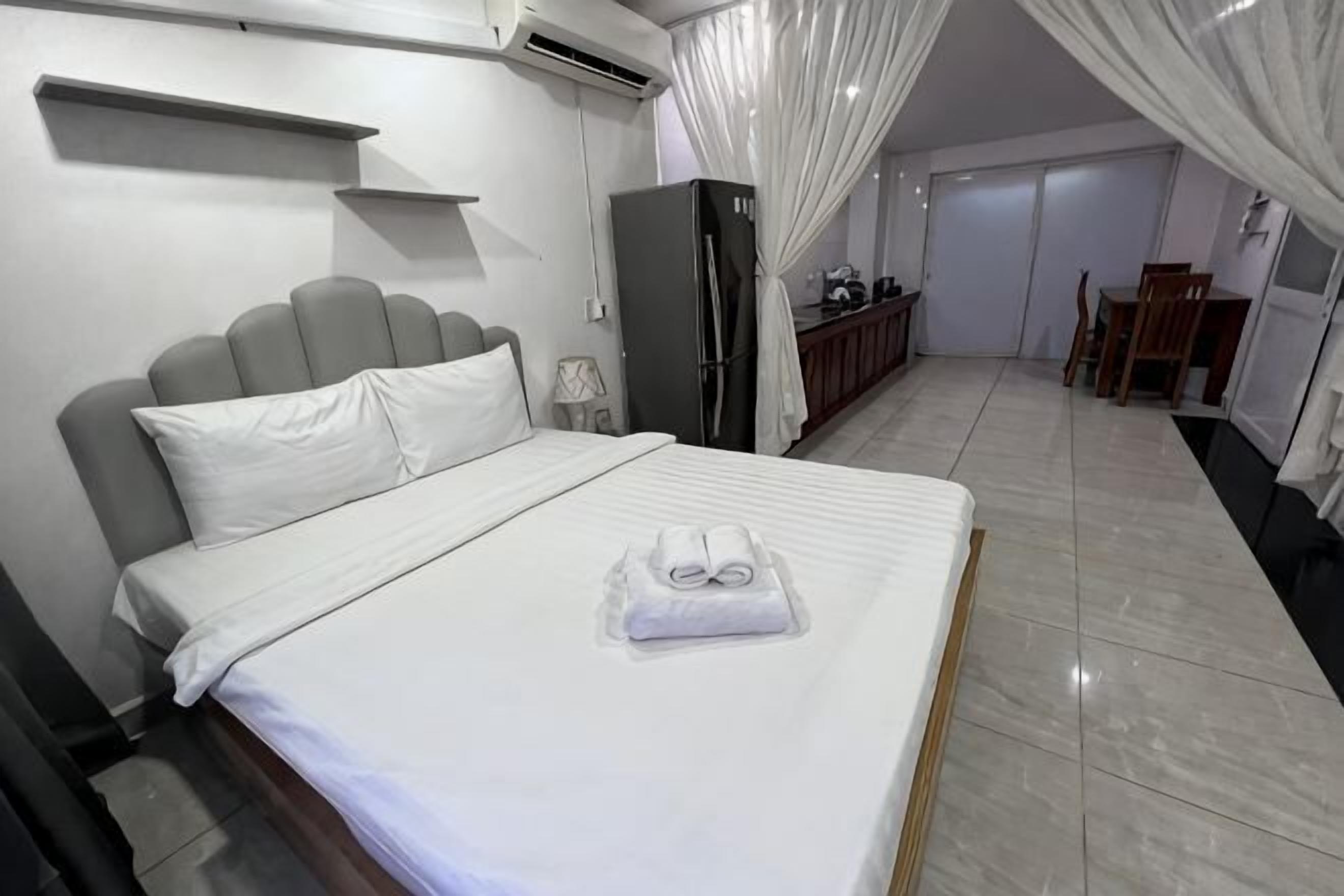 A neatly made bed with a white comforter and pillows, a grey headboard, a small bedside table with a lamp, and a wardrobe.
