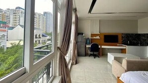 A modern hotel room with a large window, a desk with a chair, a bed, and a TV. - PINE TREE APT (Ho Chi Minh City)