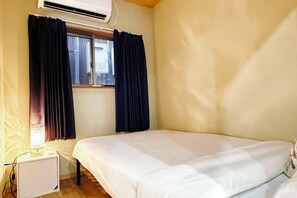 A single bed in a room with a window, a bedside table, and an air conditioning unit. - LAN A (Osaka)