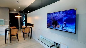 Een moderne woonkamer met een grote flatscreen televisie aan de muur, een eethoek met een glazen tafel en rieten stoelen, en een minimalistisch design met een lichtgekleurde vloer en donker plafond.
