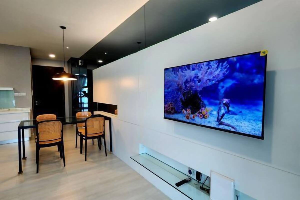 Ett modernt vardagsrum med en stor, platt-TV som är monterad på väggen, en matplats med ett glasbord och flätade stolar samt en minimalistisk design med ett ljust golv och ett mörkt tak.