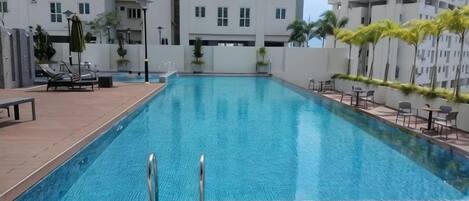 A rectangular swimming pool with a wooden deck surrounding it, set against a backdrop of multi-story residential buildings.