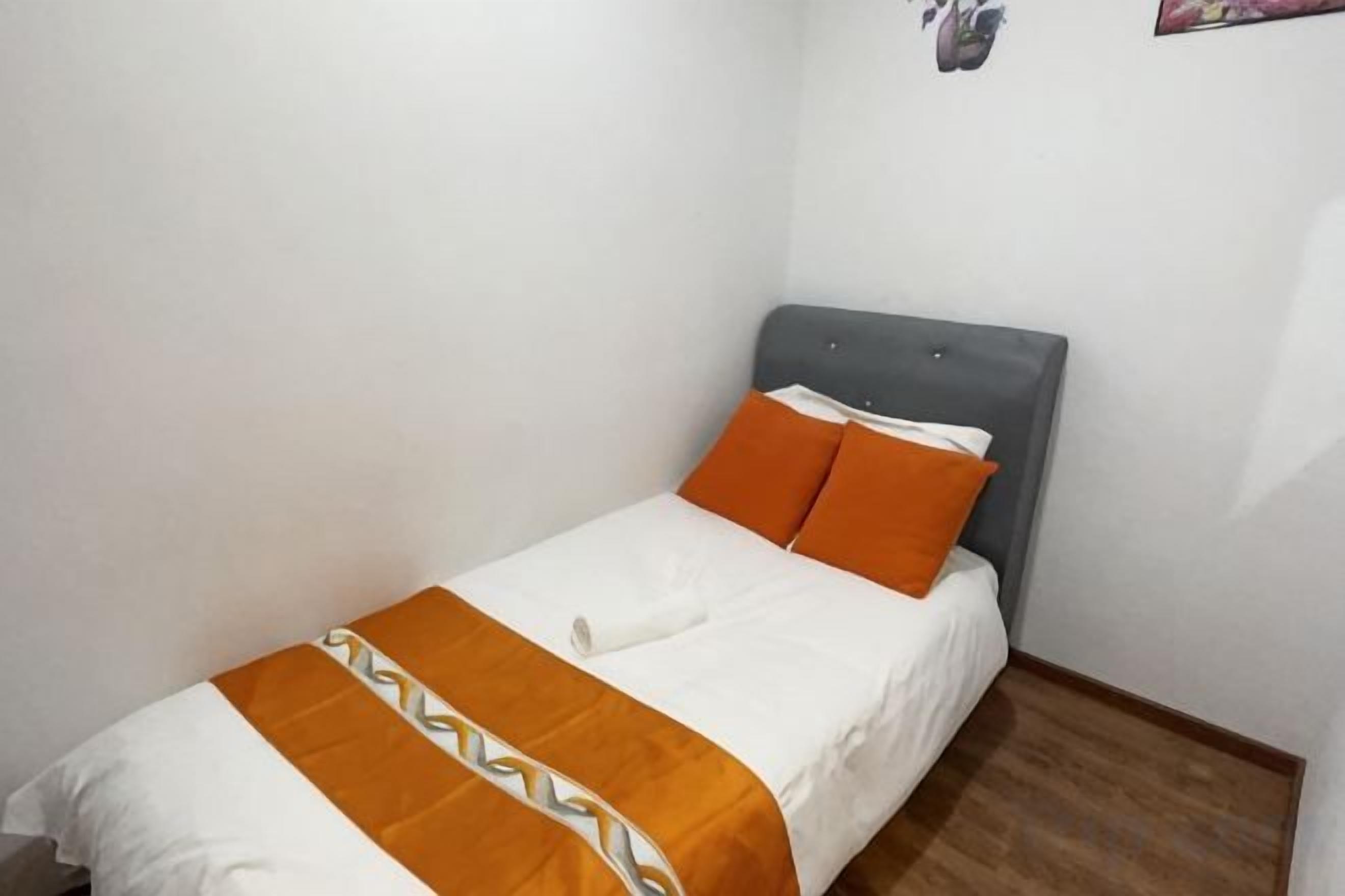 A single bed with a grey headboard, white bedding, and an orange throw blanket.