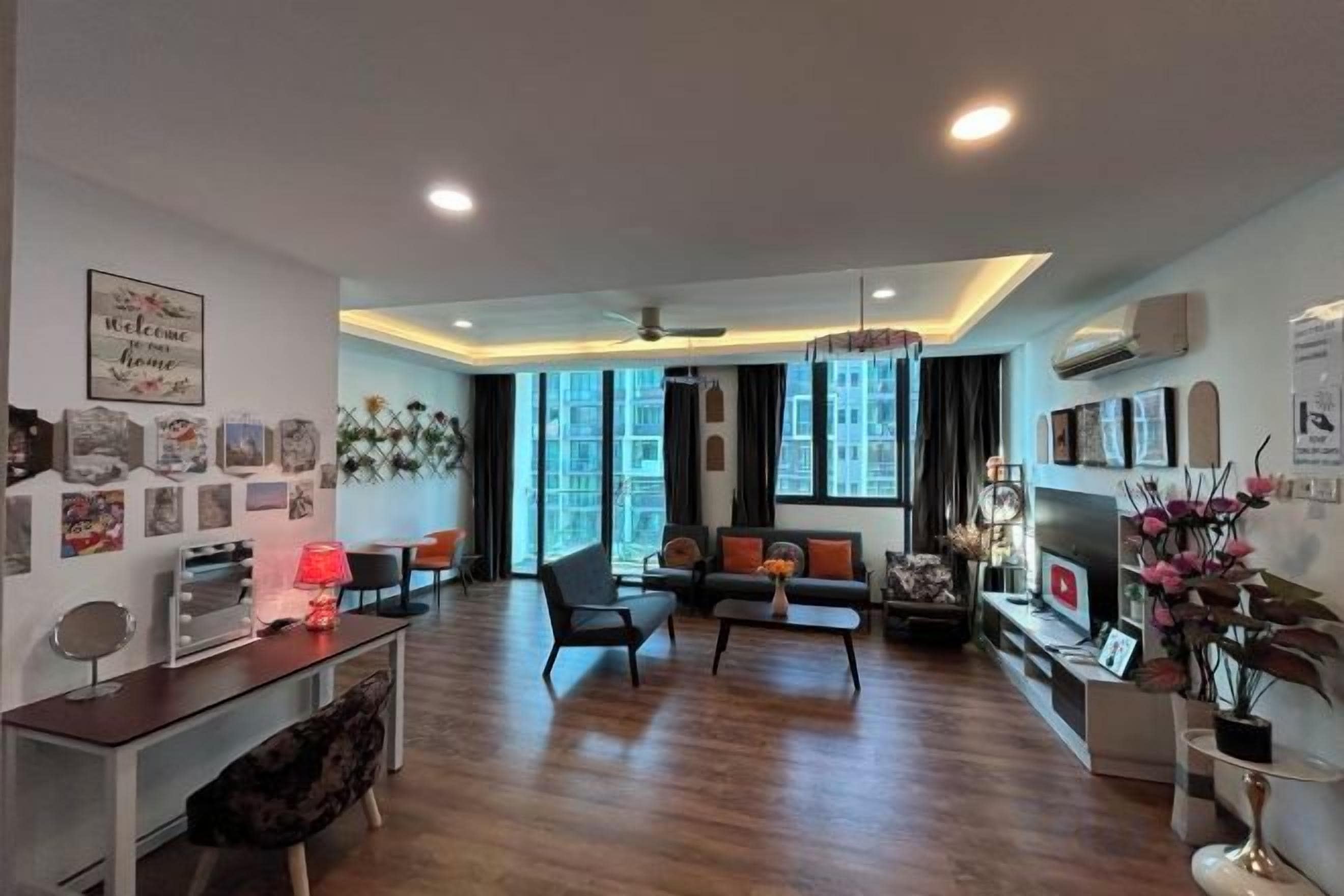 A modern living room with a sofa set, a coffee table, and a TV. There is a dining area with a table and chairs. The room has large windows and a ceiling fan.