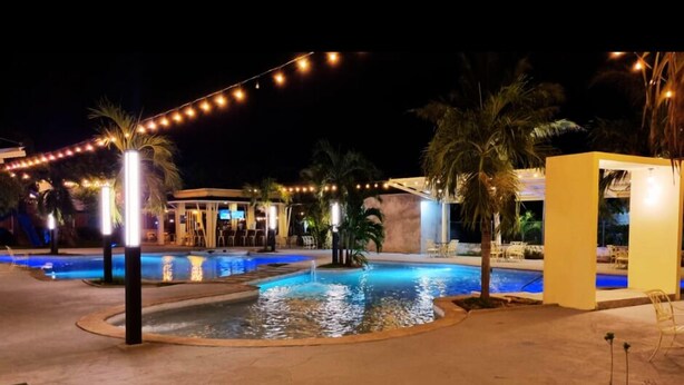 Un área de piscina nocturna con luces colgantes, palmeras y un pequeño edificio.