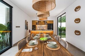 A modern dining area with a wooden table set for six, surrounded by chairs, and a kitchen in the background. - Villa Arcadia by BaliSuperHost (Bali)