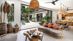 A modern living room with a curved sofa, a wooden coffee table, and a dining area with a wooden table and chairs. - Villa Arcadia by BaliSuperHost (Bali)