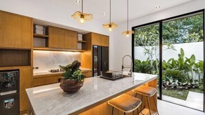 A modern kitchen with a marble countertop, wooden cabinets, a built-in refrigerator, and a large window with a view of greenery. - Villa Arcadia by BaliSuperHost (Bali)