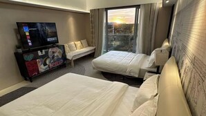 Quarto de hotel com duas camas, televisão, sofá e varanda com vista para a cidade.