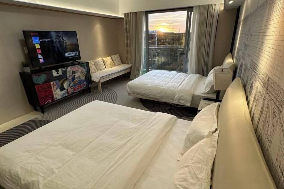 Habitación de hotel con dos camas, televisión, sofá y balcón con vista a la ciudad.