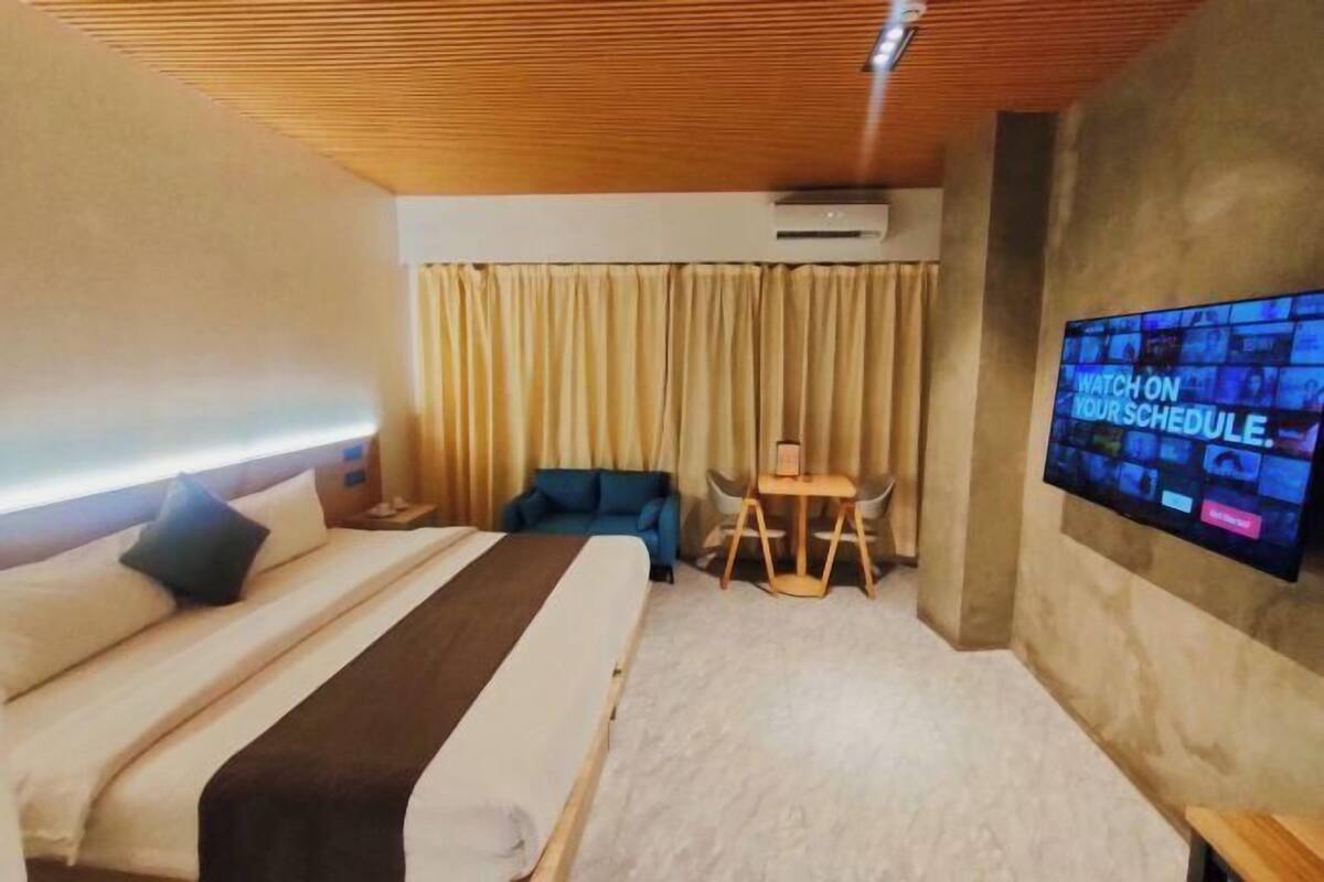 A hotel room with a bed, a TV mounted on the wall, a small table, and a blue chair.
