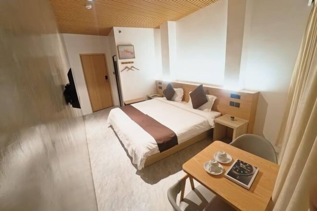 A hotel room with a bed, bedside table, and a small table with cups and a book.