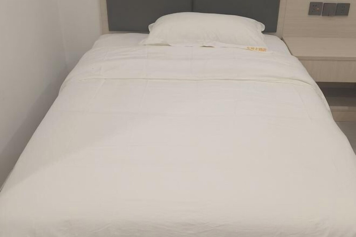 Un lit simple avec un matelas et des oreillers blancs, une tête de lit grise et un mur de couleur claire.