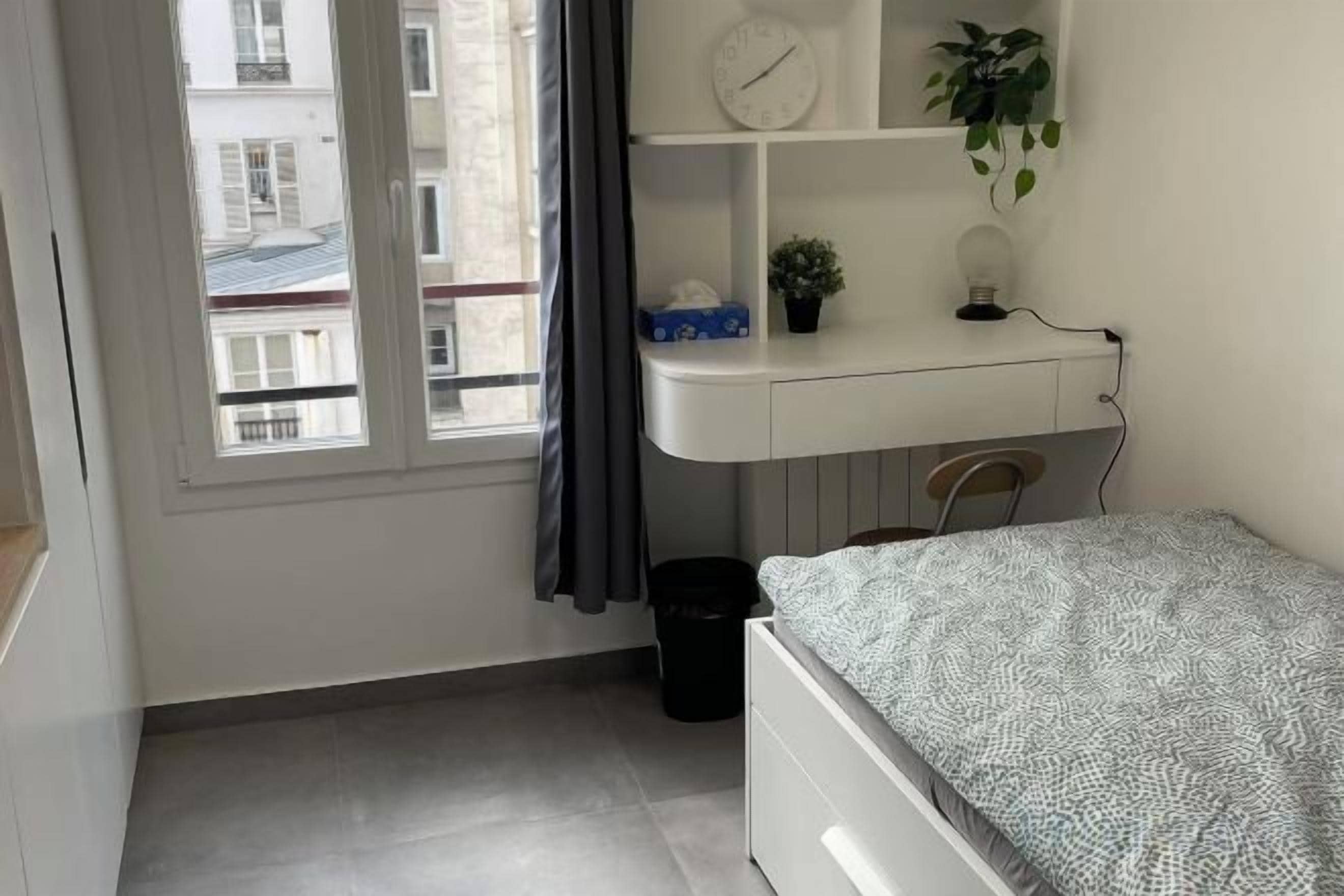 A bedroom with a bed, a desk, a chair, a window, and a plant.