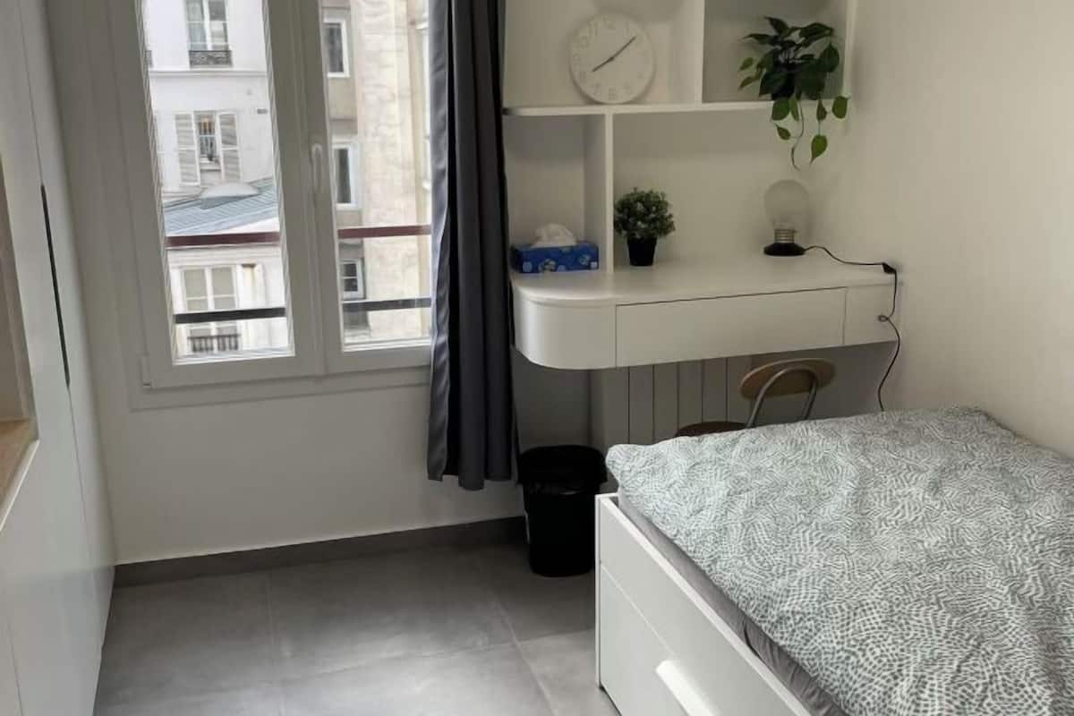 A bedroom with a bed, a desk with a clock, a potted plant, and a window with curtains.