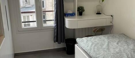 Ein Schlafzimmer mit einem Bett, einem Schreibtisch mit einer Uhr, einer Zimmerpflanze und einem Fenster mit Vorhängen.