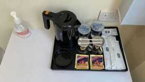 A coffee maker, a bottle of hand sanitizer, a tray with coffee cups, a spoon, a napkin, and two packets of coffee creamer on a white table.