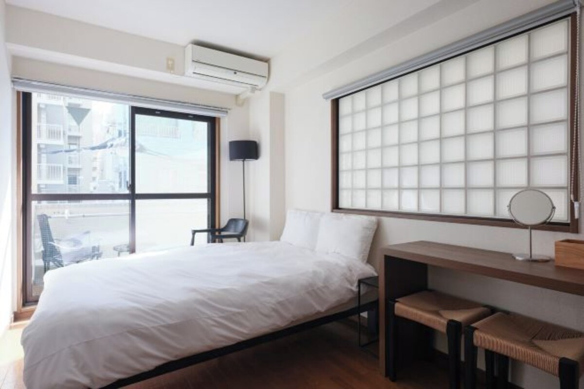 A modern hotel room with a large bed, a desk with a chair, a small table, and a large window with a view of buildings.