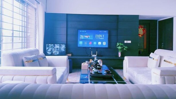 Un salon moderne avec un grand téléviseur à écran plat fixé sur un mur bleu foncé, deux canapés blancs, une table basse sur laquelle sont disposés divers objets, et une fenêtre avec des stores.