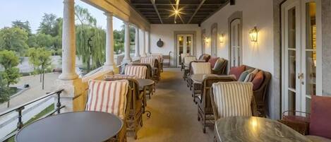 A covered outdoor seating area with wicker furniture and tables, a view of the outdoors, and a stone wall.
