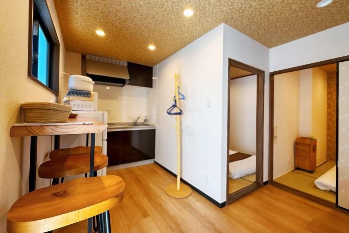 A compact hotel room with a kitchenette, a wooden bench, and a mirror reflecting the room.