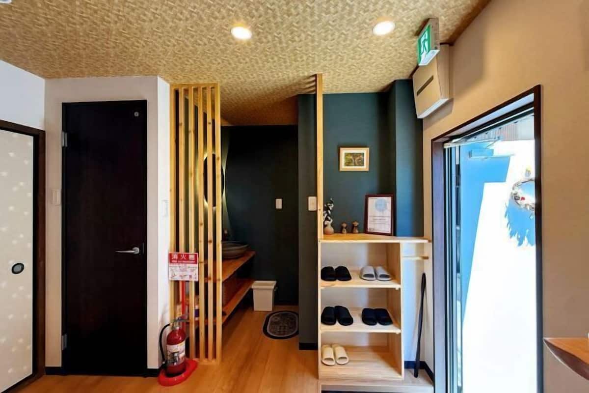 A hotel room with a wooden floor, a black door, a fire extinguisher, a shoe rack, and a window with a view of the outside.