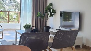 A modern living room with a TV, two chairs, a small table, a potted plant, and a view of trees through a balcony. - RR Homestay D' Awana Kijal (Kijal)