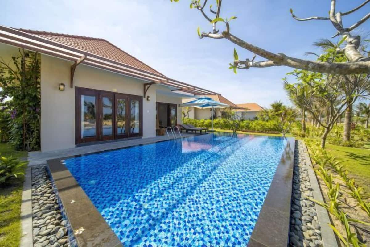A swimming pool with clear blue water, surrounded by a stone border and lush greenery.