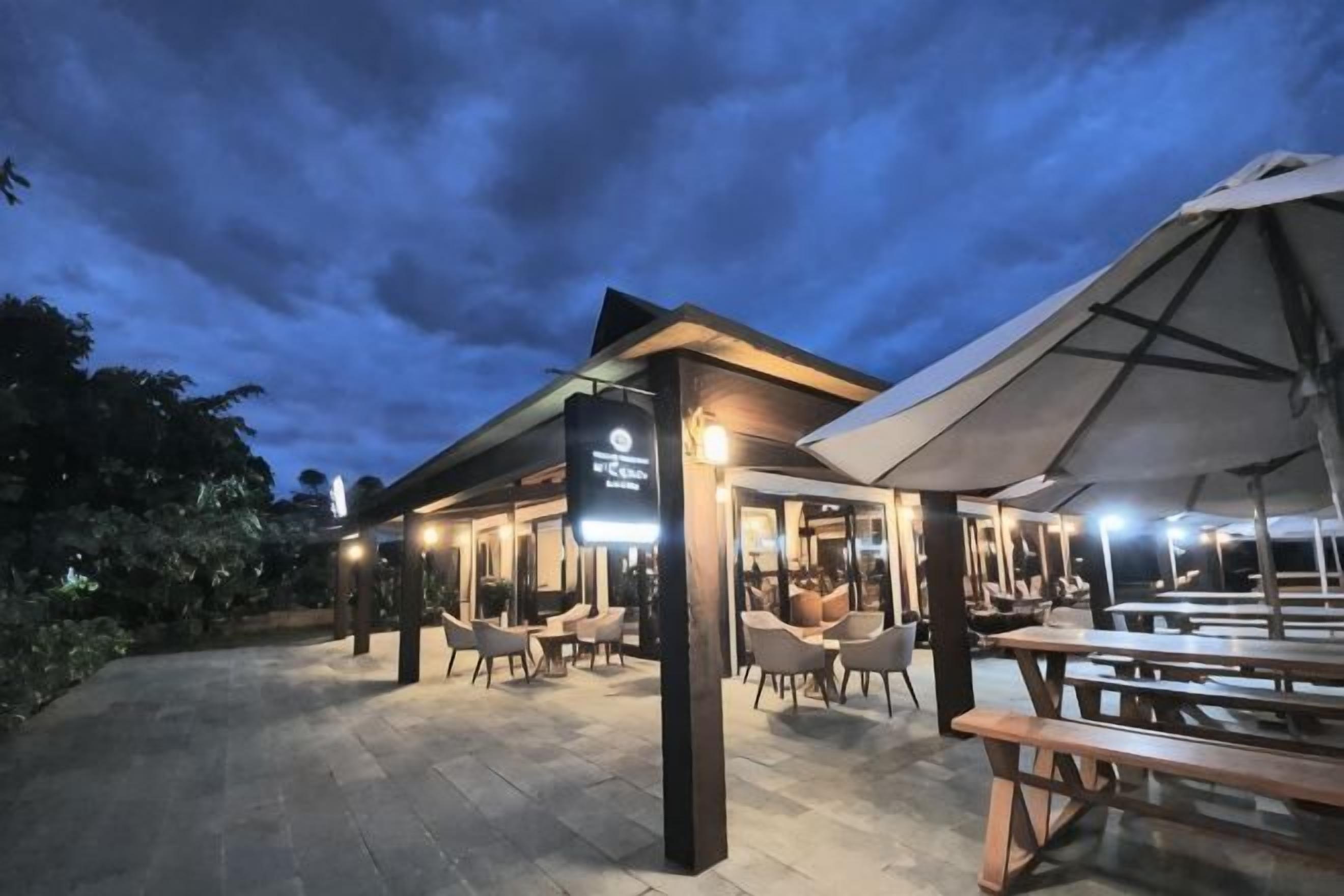 Área externa com mesas e cadeiras, parte coberta e sinalização indicando restaurante ou espaço de refeições.
