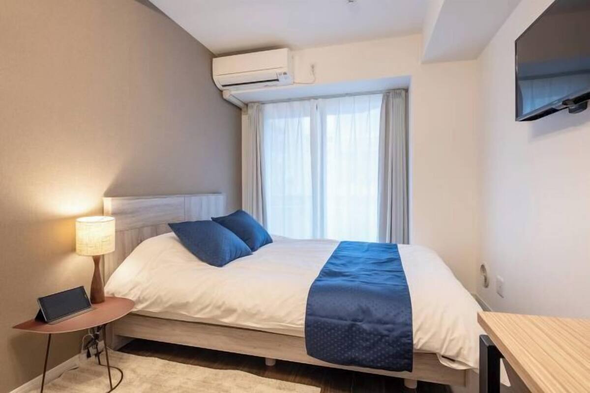 A hotel room with a bed, a bedside table with a lamp, a TV mounted on the wall, and an air conditioning unit above the window.