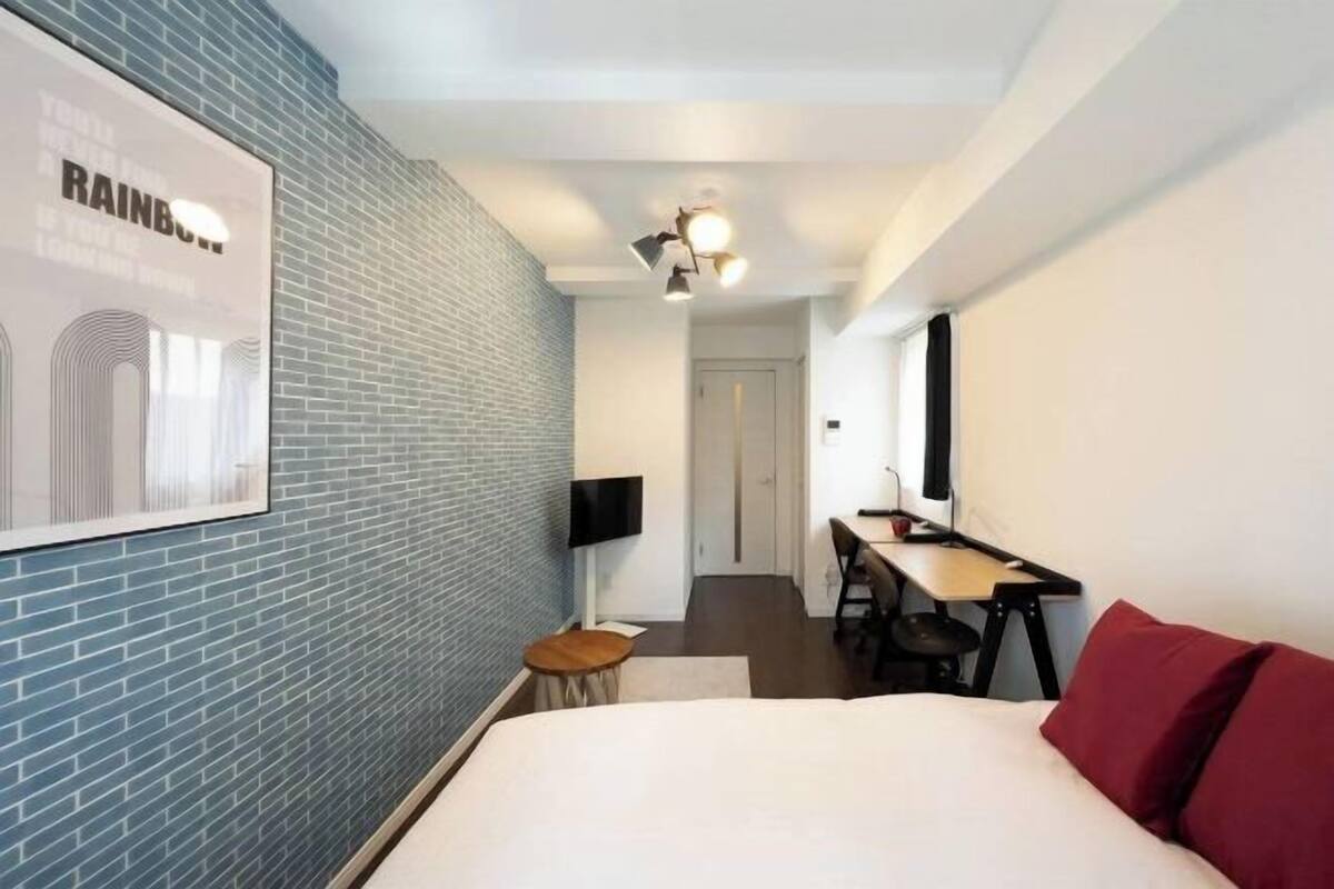 A hotel room with a brick wall, a bed with red pillows, a desk with a chair, a TV, and a ceiling light fixture.