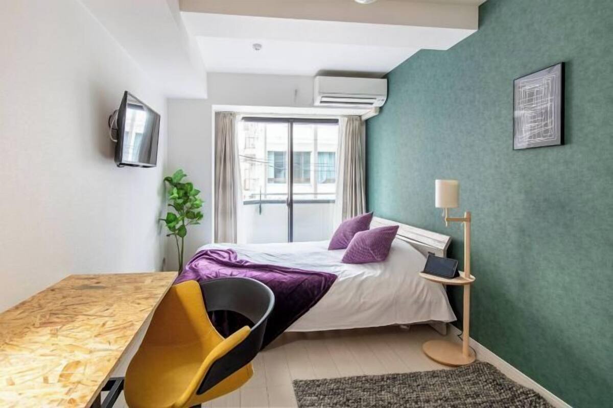 A modern bedroom with a bed, a desk, a chair, a plant, and a window.