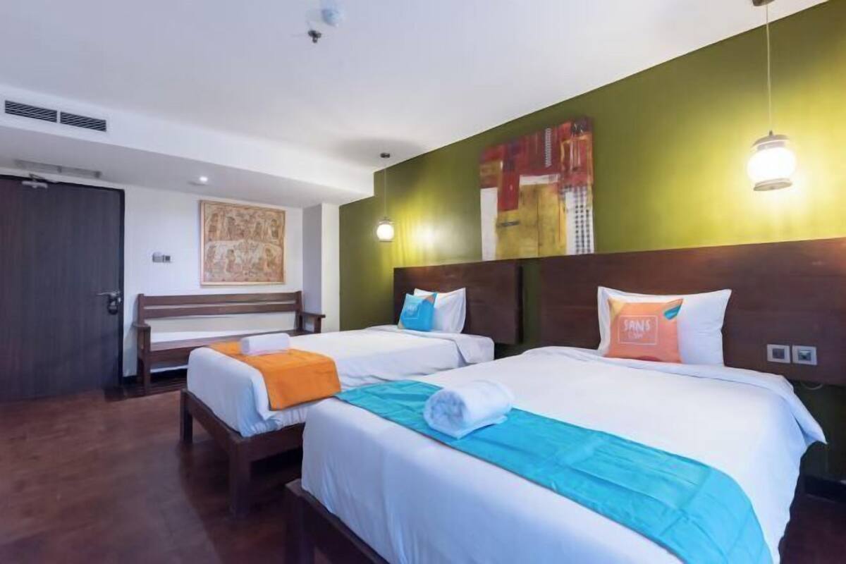A hotel room with two beds, a bench, a green accent wall, and a painting.