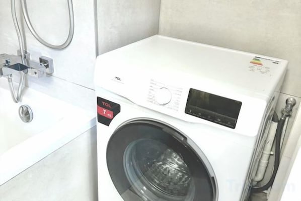A front-loading washing machine with a control panel, a water inlet hose, and a metal pipe. - Apartement Choisy (Paris)