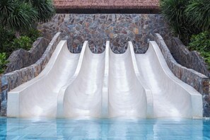 A pool with three water slides against a stone wall backdrop. - Apec Mandala Chambay Mui Ne - Oceanic Studio (Phan Thiet)