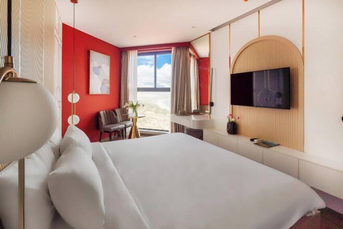 Ein Hotelzimmer mit einem großen Bett, einem Schreibtisch, einem Stuhl, einem an der Wand montierten Fernseher und einem Fenster mit Vorhängen.