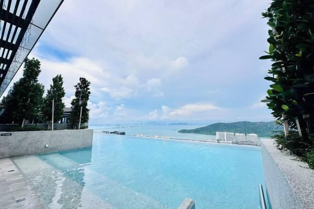 A swimming pool with clear blue water, surrounded by a stone wall and greenery, with a view of the sea and distant hills.