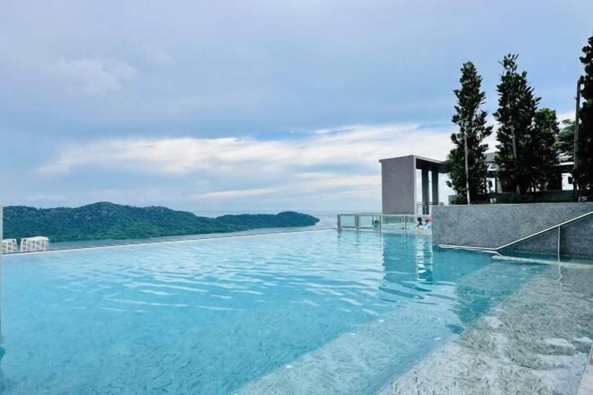 A clear blue infinity pool with a view of a distant island, surrounded by a modern building and tall trees.