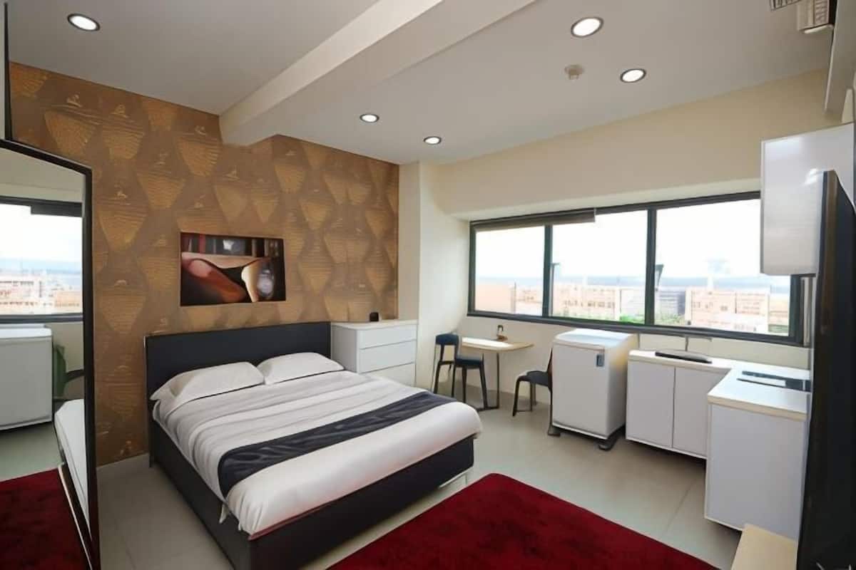 A modern hotel room with a large bed, a desk, a chair, a small refrigerator, and a large window offering a city view.