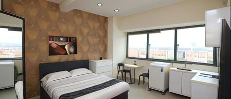 A modern hotel room with a large bed, a desk, a chair, a small refrigerator, and a large window offering a city view.